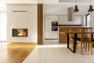 Modern kitchen and living room area with an open floor plan. A table and chairs are set up in the foreground and a fire is lit in a white stone fireplace in the background.