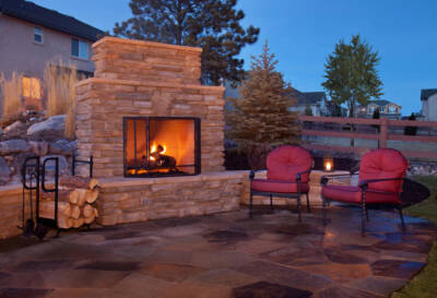 Evening shot of a backyard patio area featuring a stone fireplace with a fire lit inside. Two red arm chairs are set up to the right of the fireplace.
