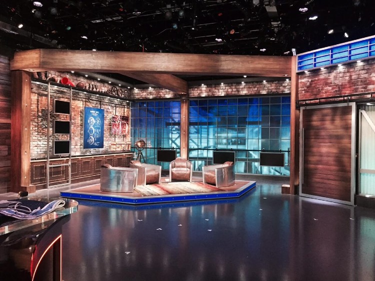 Overview of a television studio, featuring natural wood and leather seats set against glass and red brick faux wall panels.