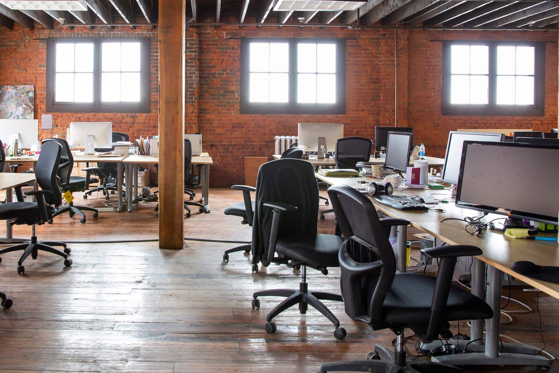 Work stations within a large, industrial style open office space featuring brick walls, wood panel floors and wooden beams.