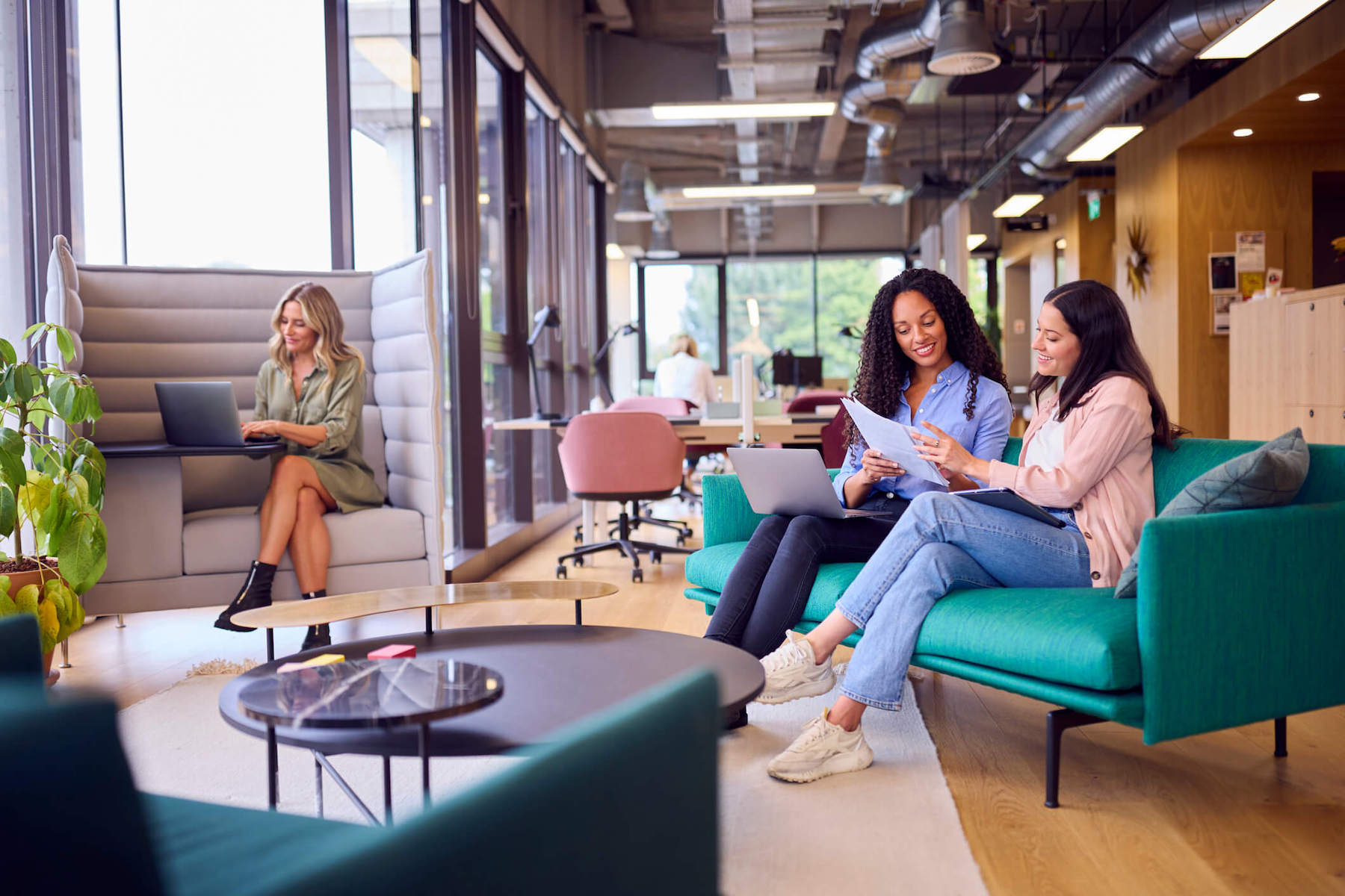 Two women having a meeting in an open common area, while another works in an open booth.