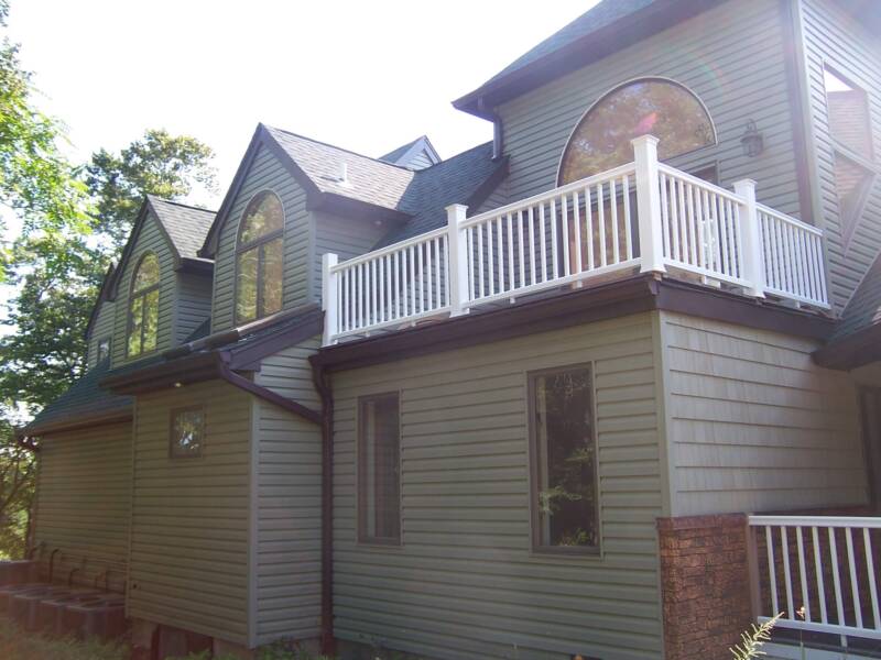 Backside of a large family home, with dark green siding and shingles, white fencing and a half wall made of brown stone faux wall panels.