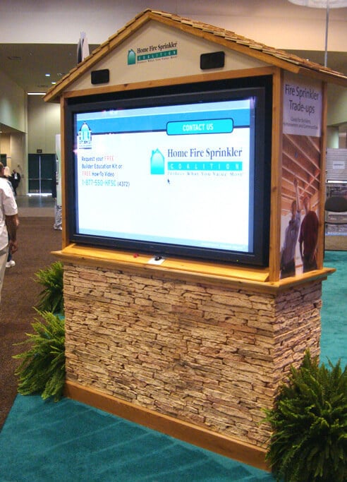 A stand alone trade show display with a wood shingled roof and faux stone panels, holding a monitor that displays information for the Home Fire Sprinkler Coalition.