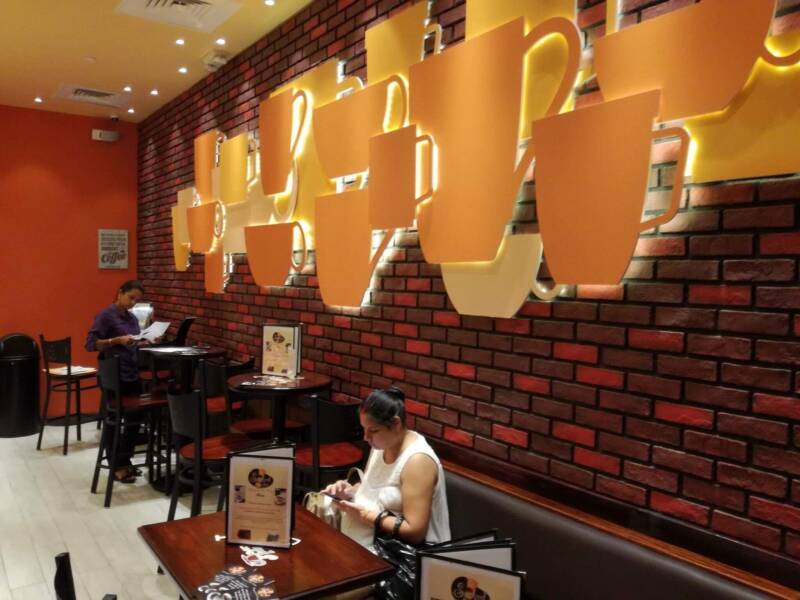 A cafe seating area featuring booths and tall tables. One woman stands beside a table and another sits at a booth in front of a brick wall decorated with yellow coffee cup accents.