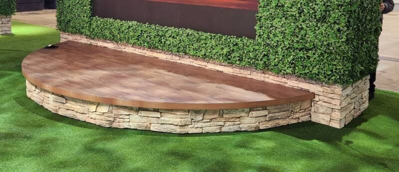 Outdoor patio area featuring a raised stage made from faux stone and wood panels. An ivy-covered backdrop accents the stage.